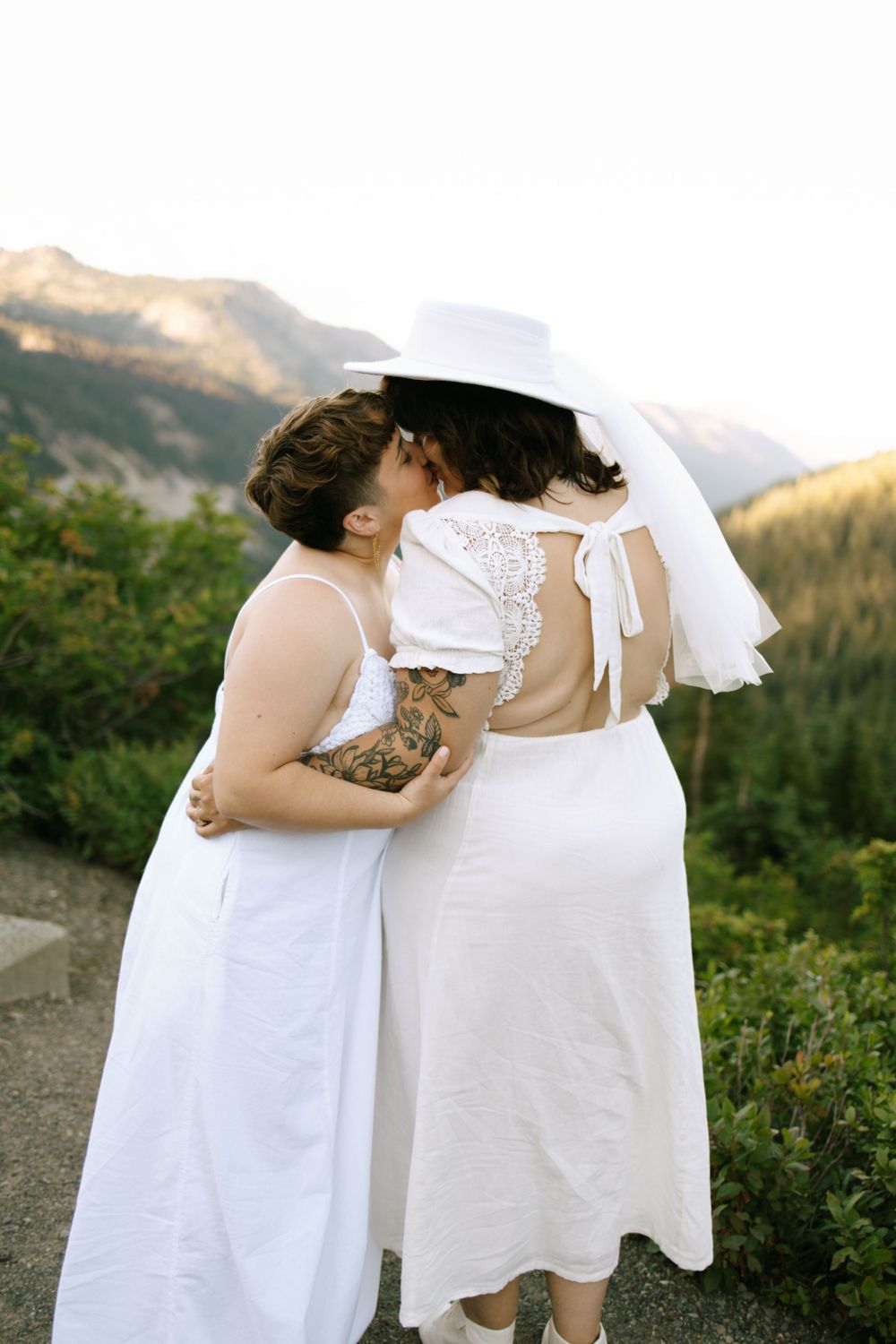 Two brides in white dresses share a kiss with mountain scenery behind them.