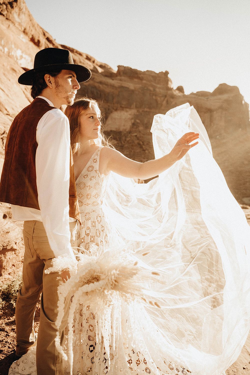 Boho couple in desert setting as bride tosses flowing veil in golden sunset light.