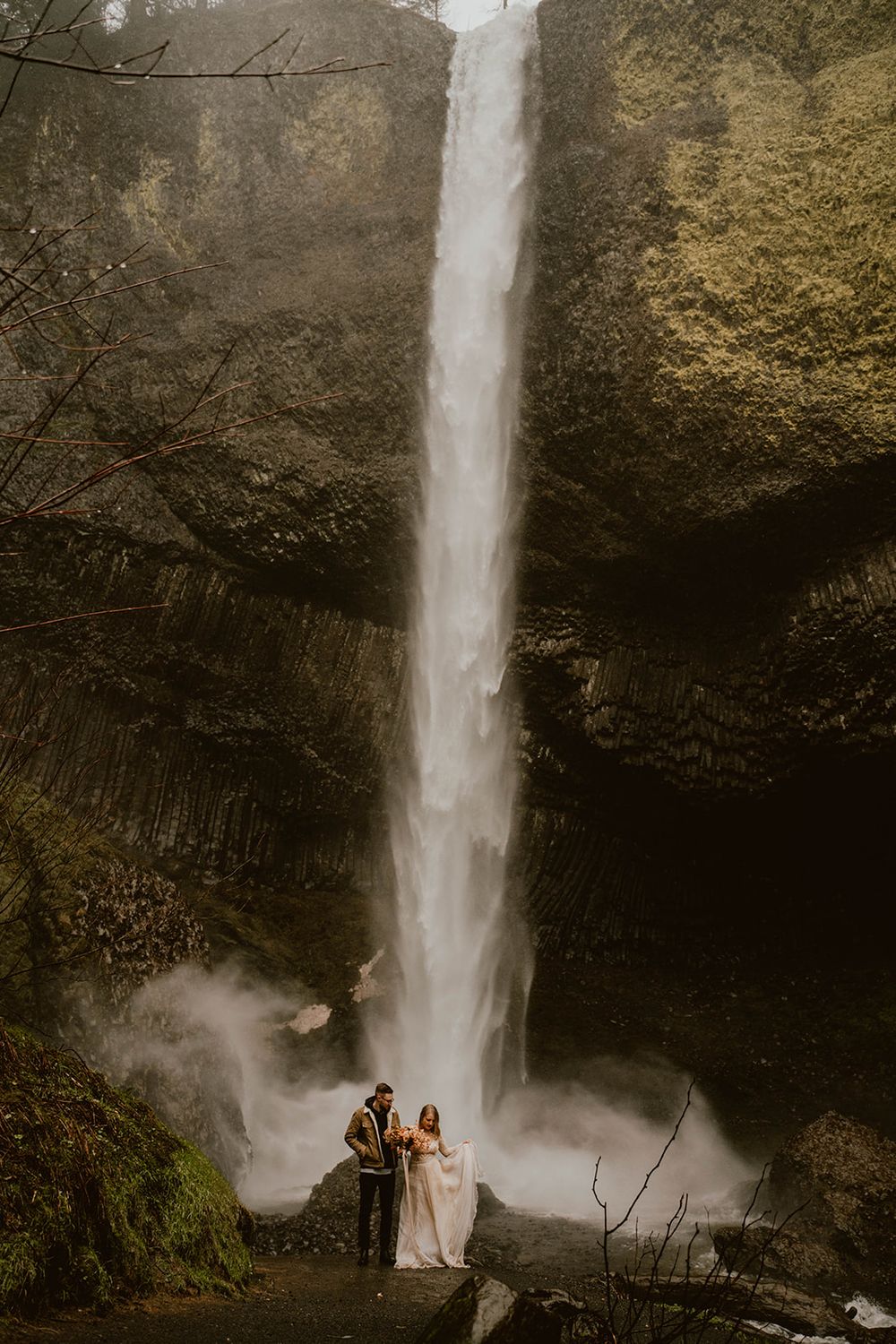 Couple elopes in front of tall waterfall in lush green forest setting.