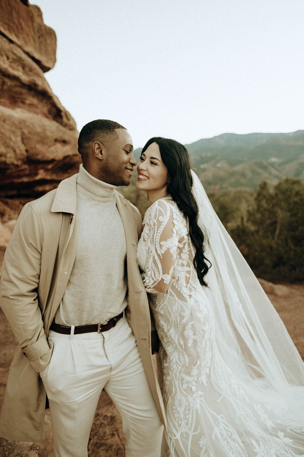 Bride in lace gown and groom in beige coat share romantic moment at Red Rocks Colorado.