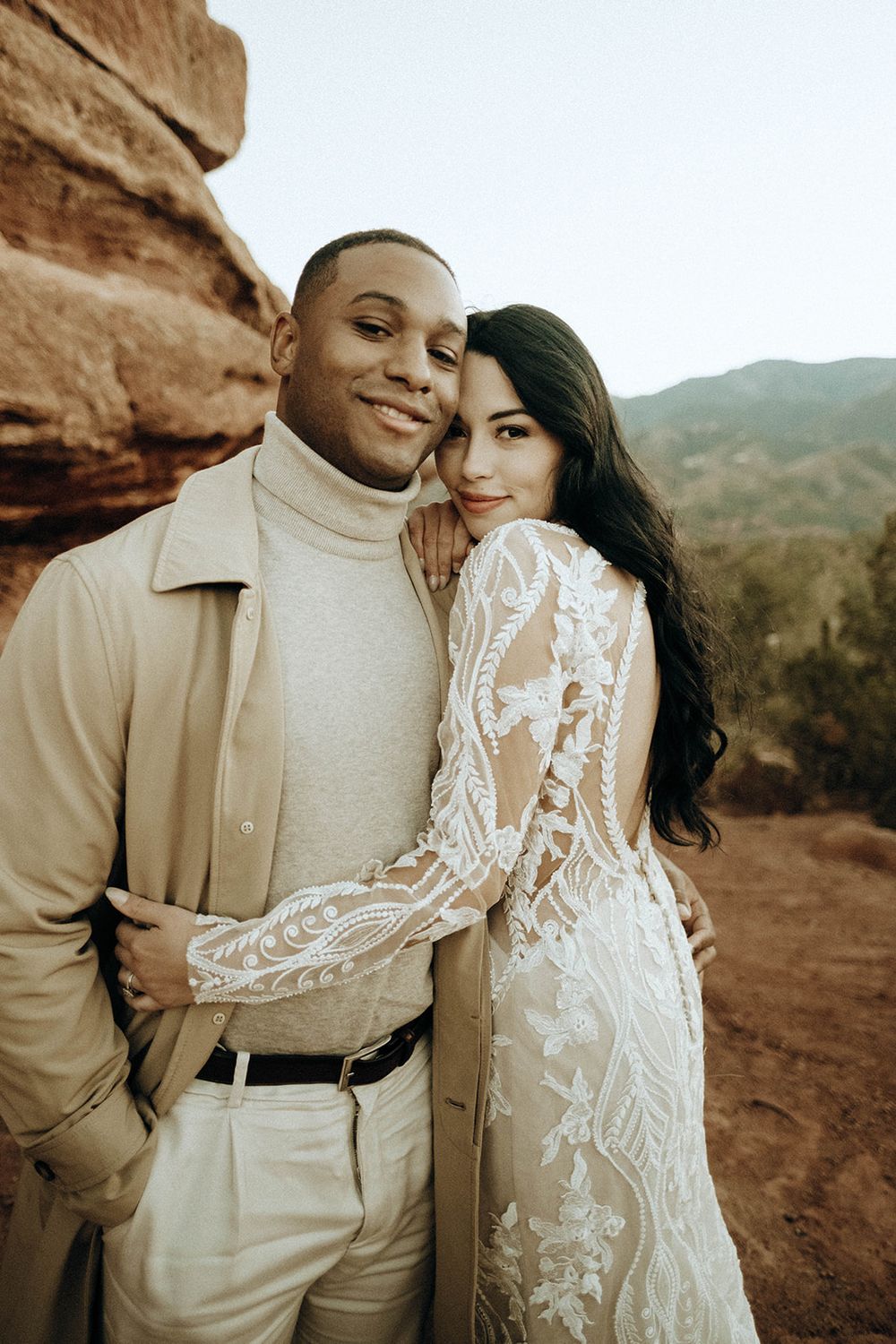 Bride in long-sleeve lace gown embraces groom in beige turtleneck at Red Rocks Colorado wedding.