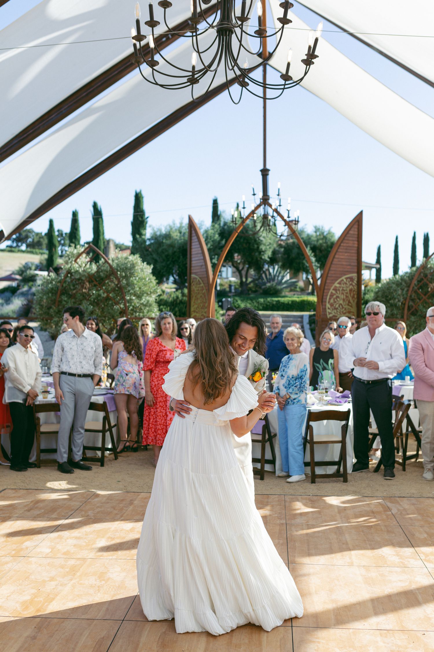 Reception scene under tent with chandelier and guests watching first dance.