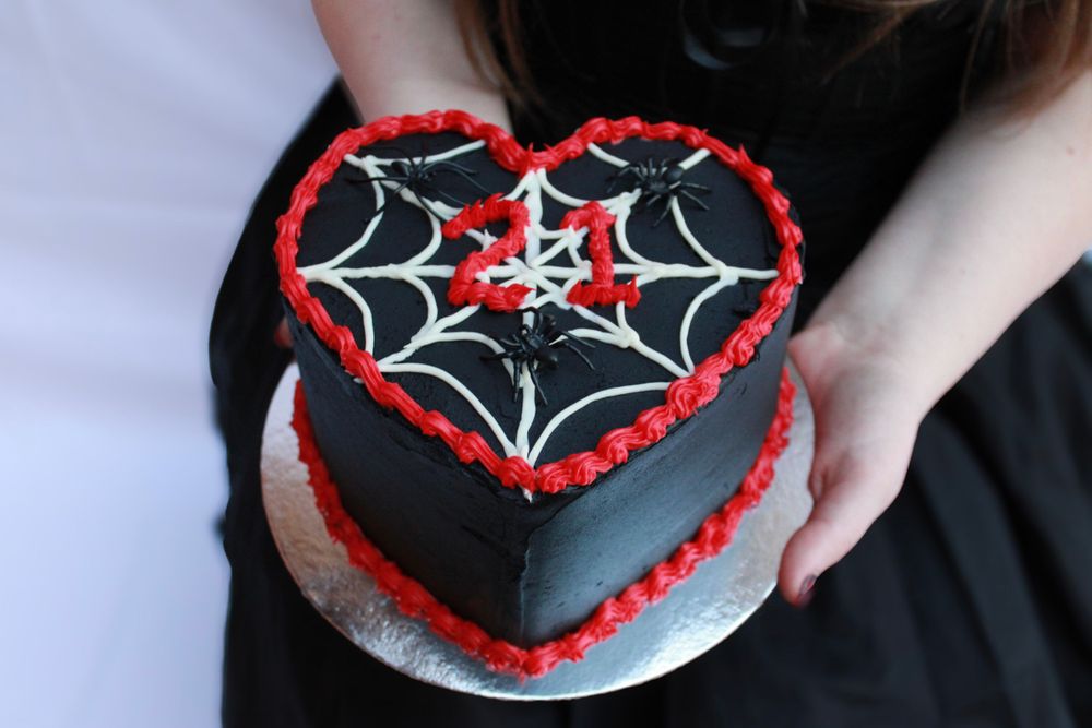 A heart-shaped black cake decorated with spider web designs and red piping.