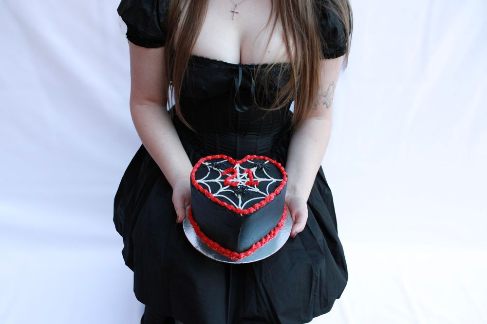 A gothic-themed black heart cake with spiderweb design being held against a dark dress.