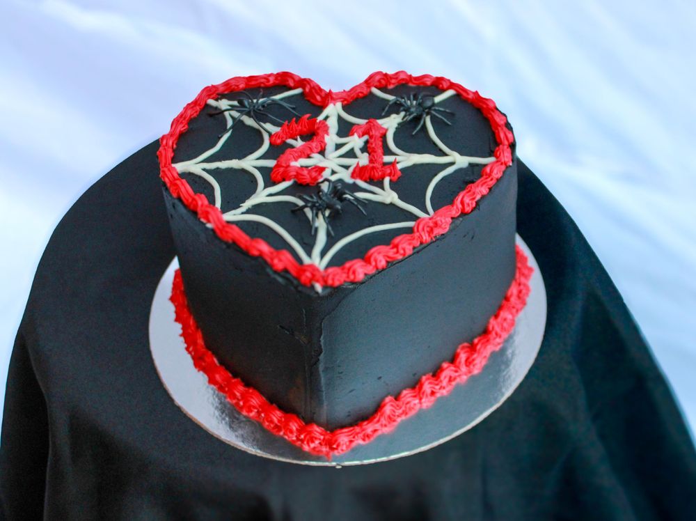 A black heart-shaped cake decorated with white spiderweb pattern and red trim on a silver cake board.