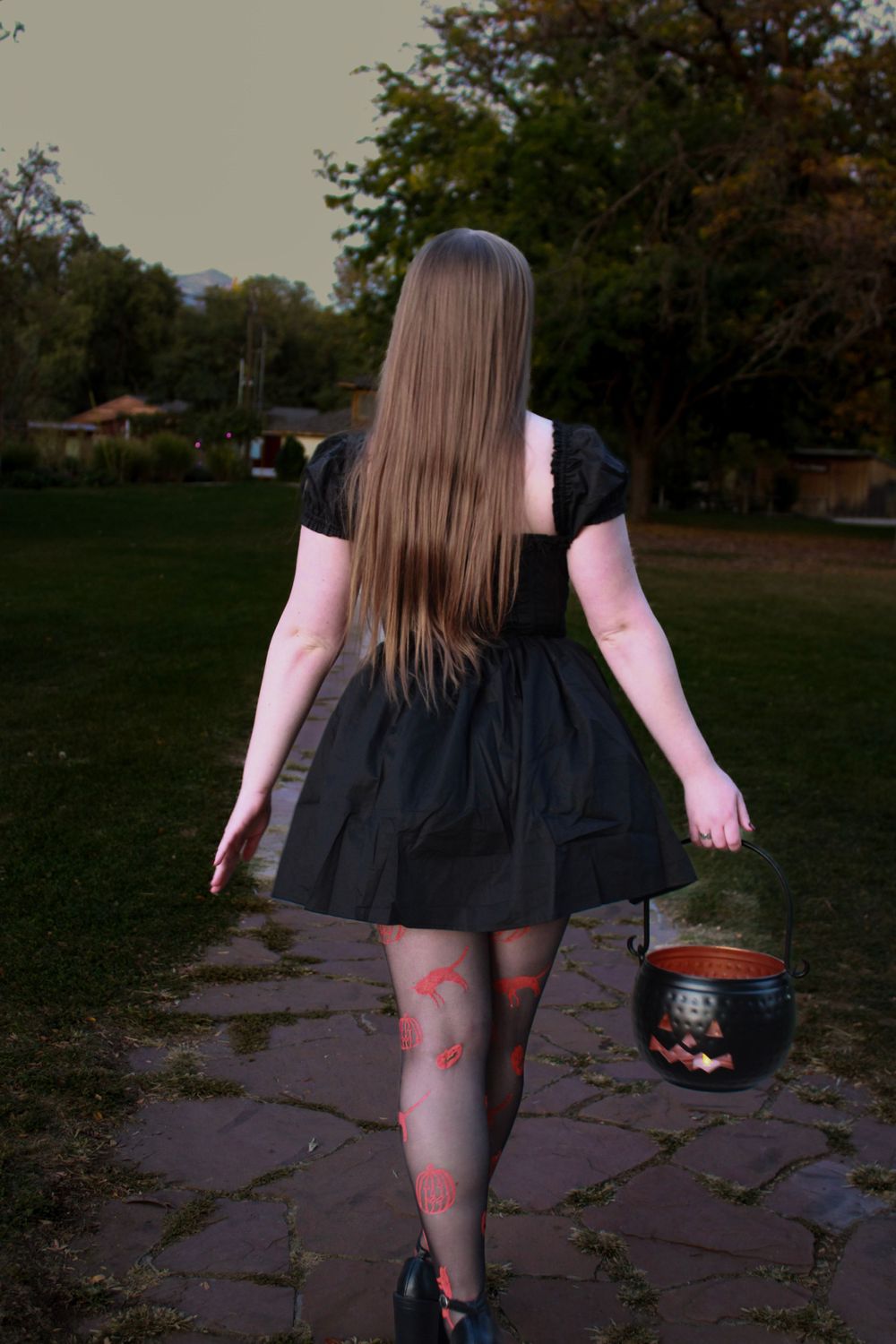 Person in black dress and patterned tights carries Halloween cauldron on stone path at dusk.