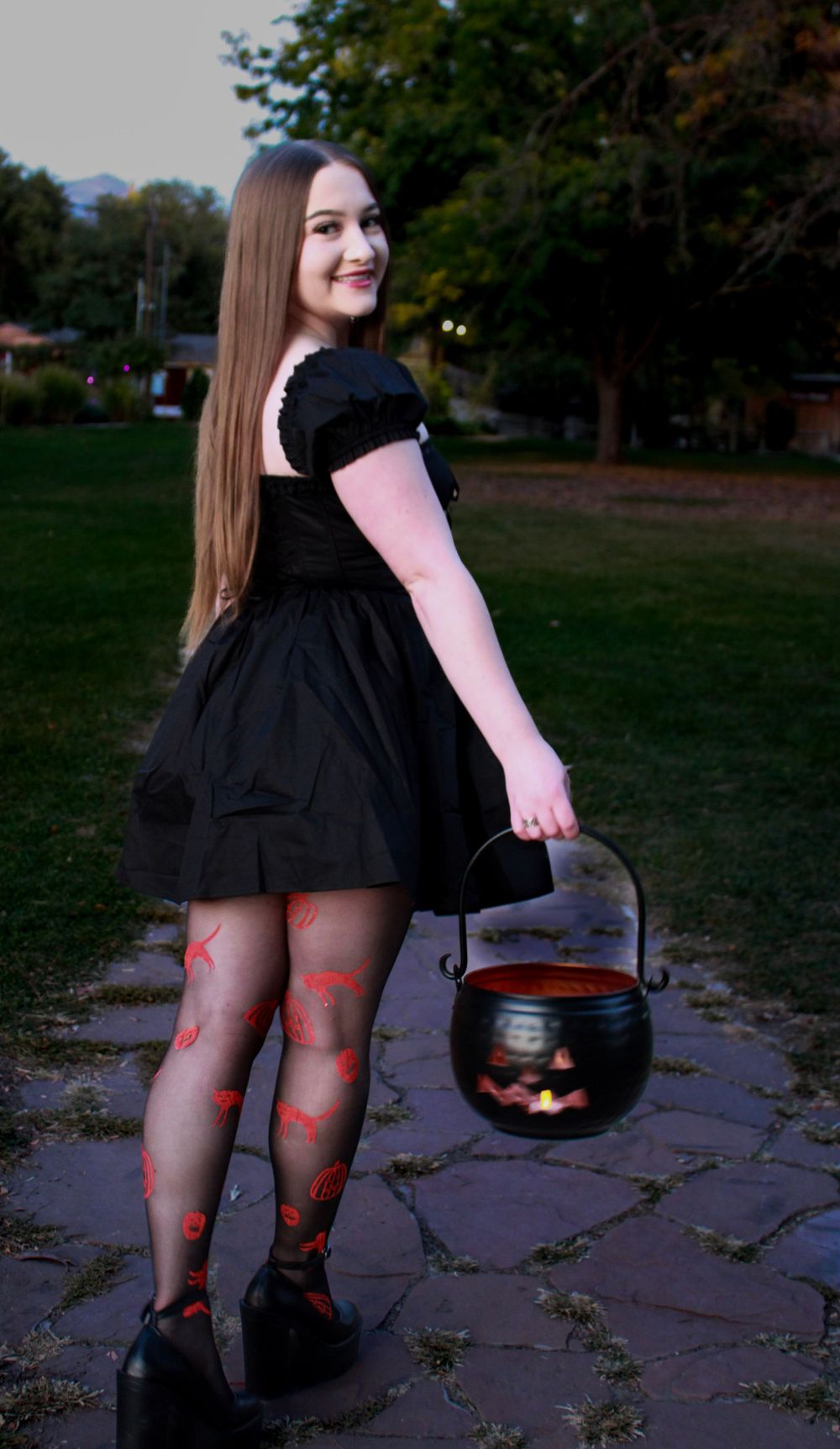 Figure in black dress holds decorative Halloween cauldron while posing outdoors.
