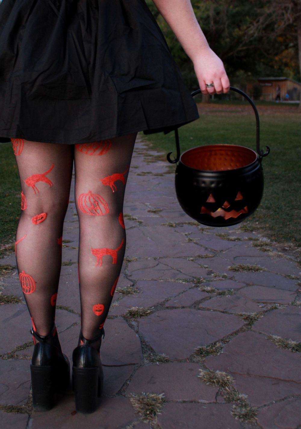 Close-up of patterned tights and black shoes with Halloween cauldron on stone pathway.