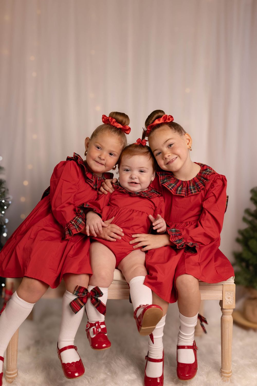 Three siblings in matching red holiday dresses pose on wooden bench.