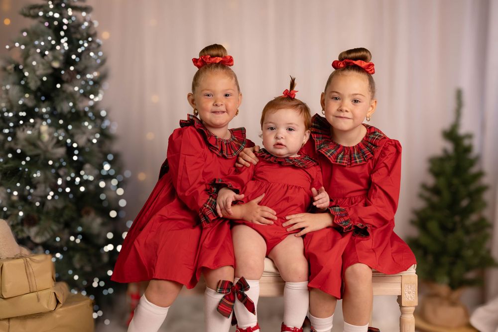 Three young girls in matching red dresses pose together by a Christmas tree.