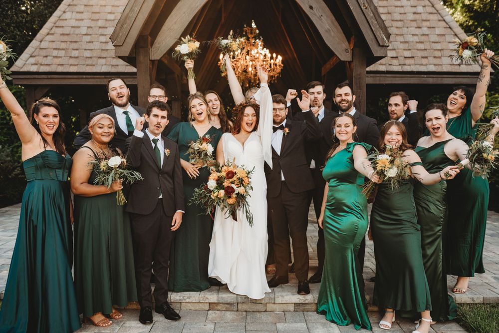 Wedding party celebrates outside chapel with bridesmaids in emerald green dresses and groomsmen in black suits.