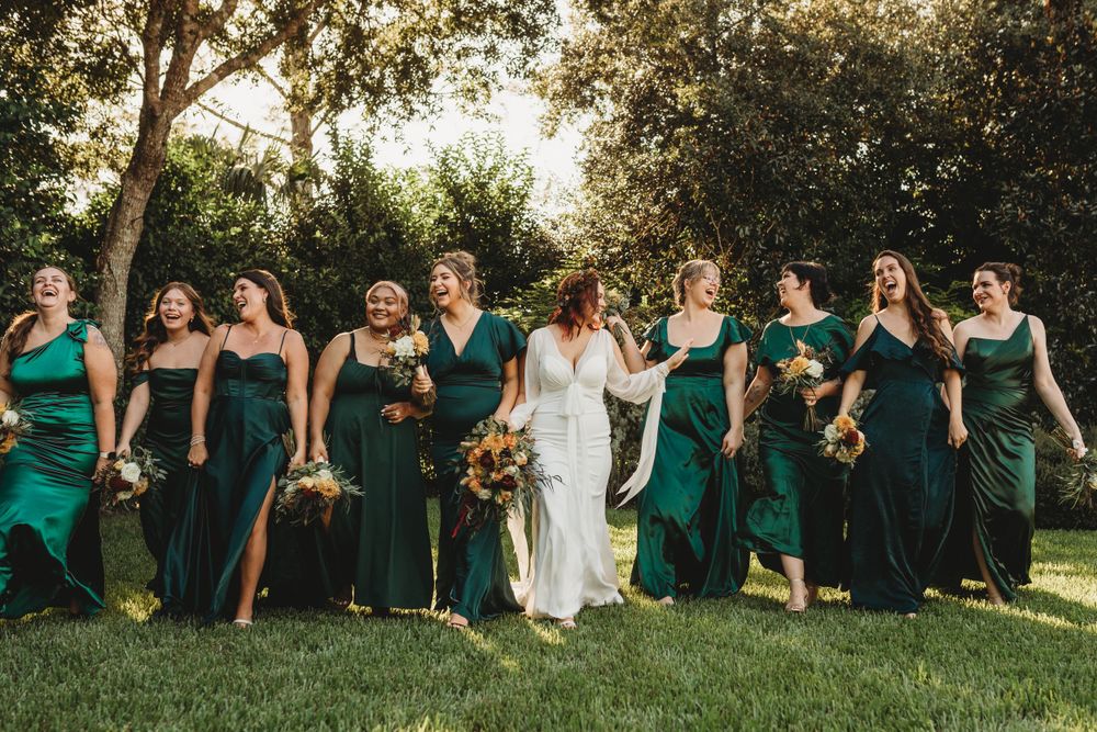 Wedding party wearing emerald green dresses walking together on sunlit grass.