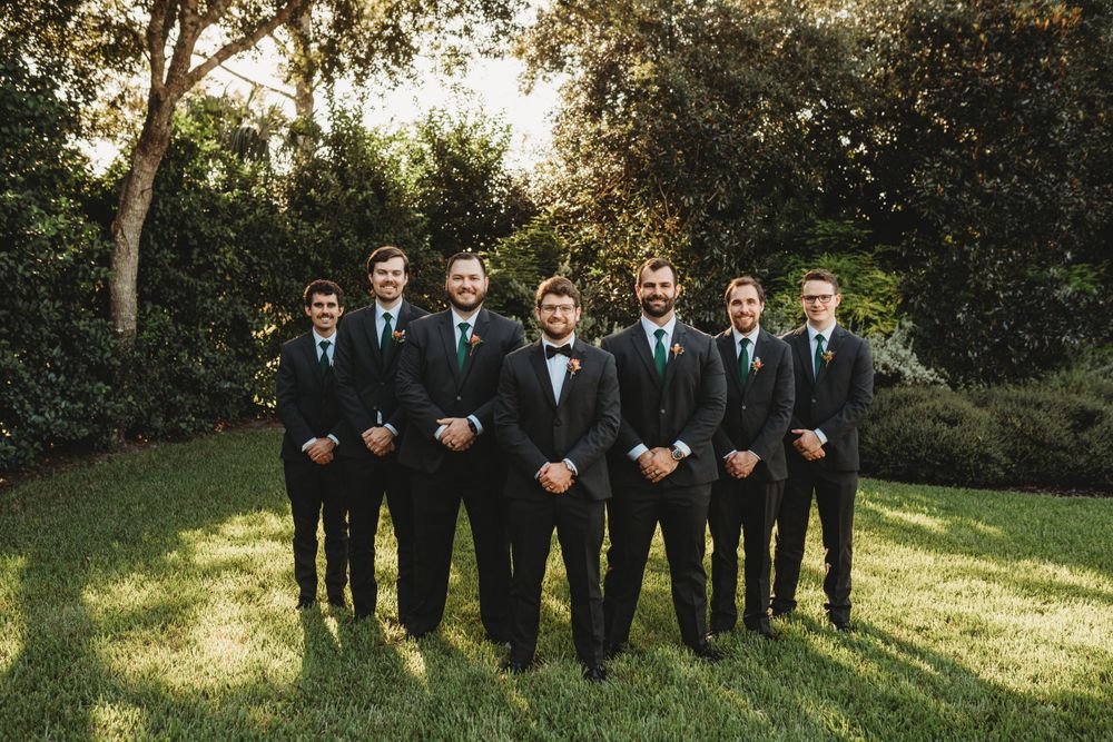 Groomsmen in black suits and blue ties pose formally on garden lawn.