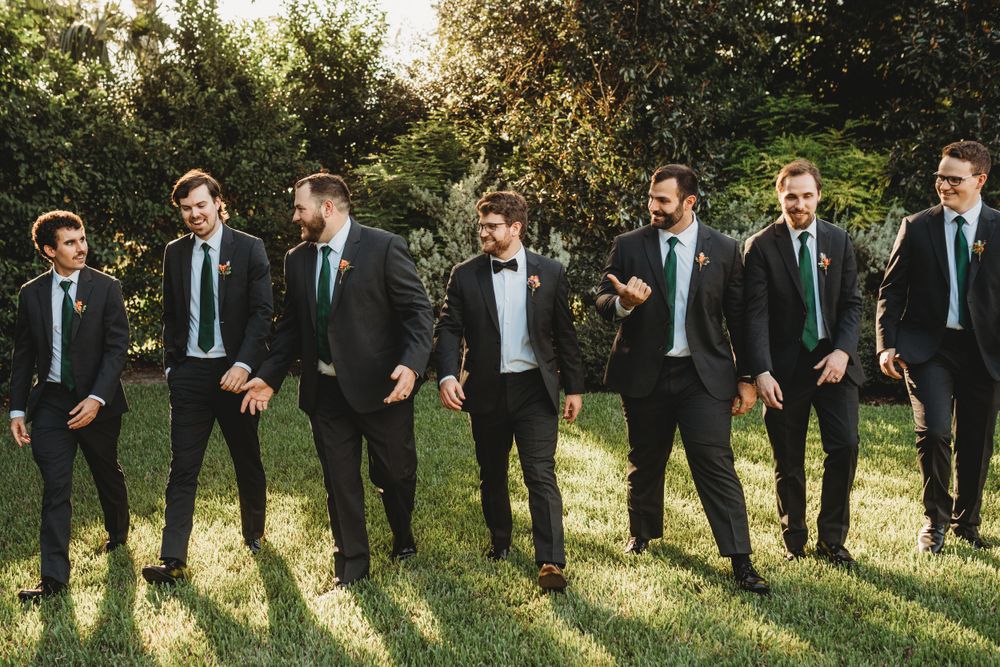 Groomsmen in dark suits share casual moment while posing outdoors.