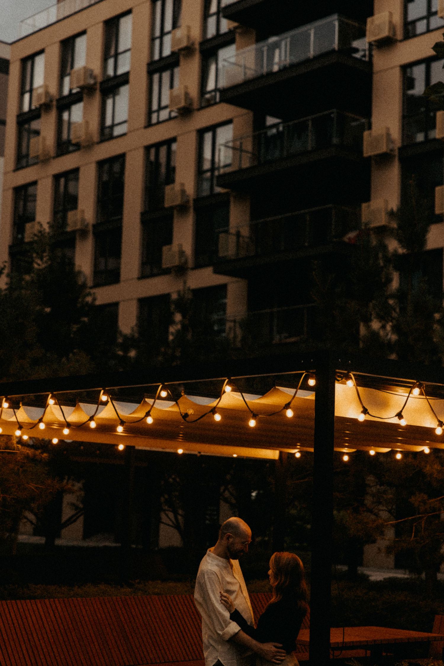 A couple stands beneath glowing string lights with an urban apartment building backdrop.
