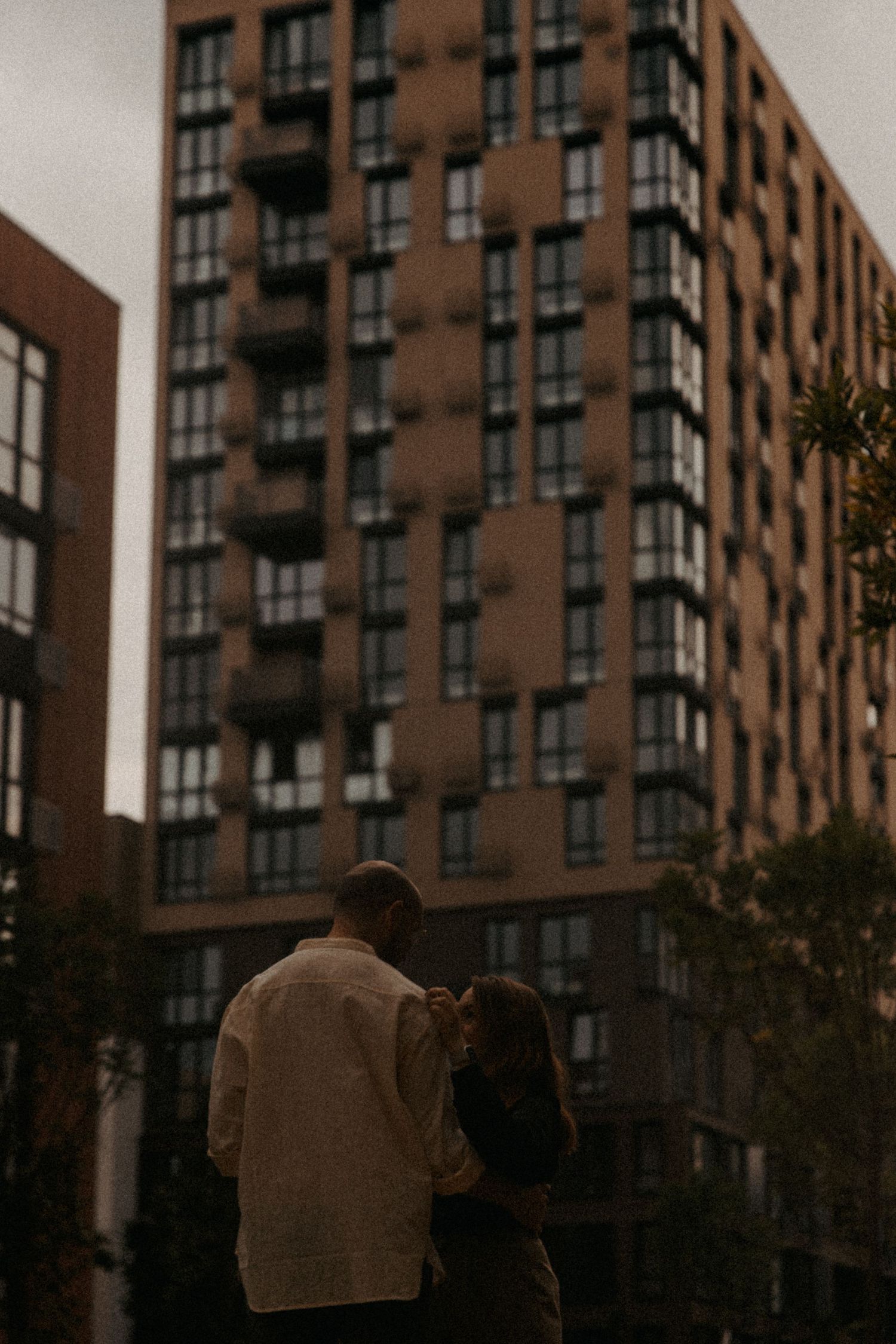 A couple stands in silhouette against a tall apartment building at dusk.