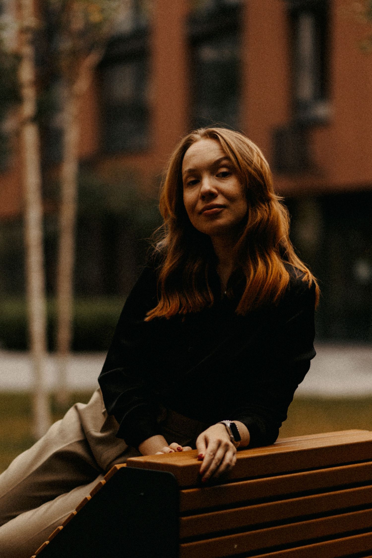 Person in black sweater sits on wooden bench with brick building in background.