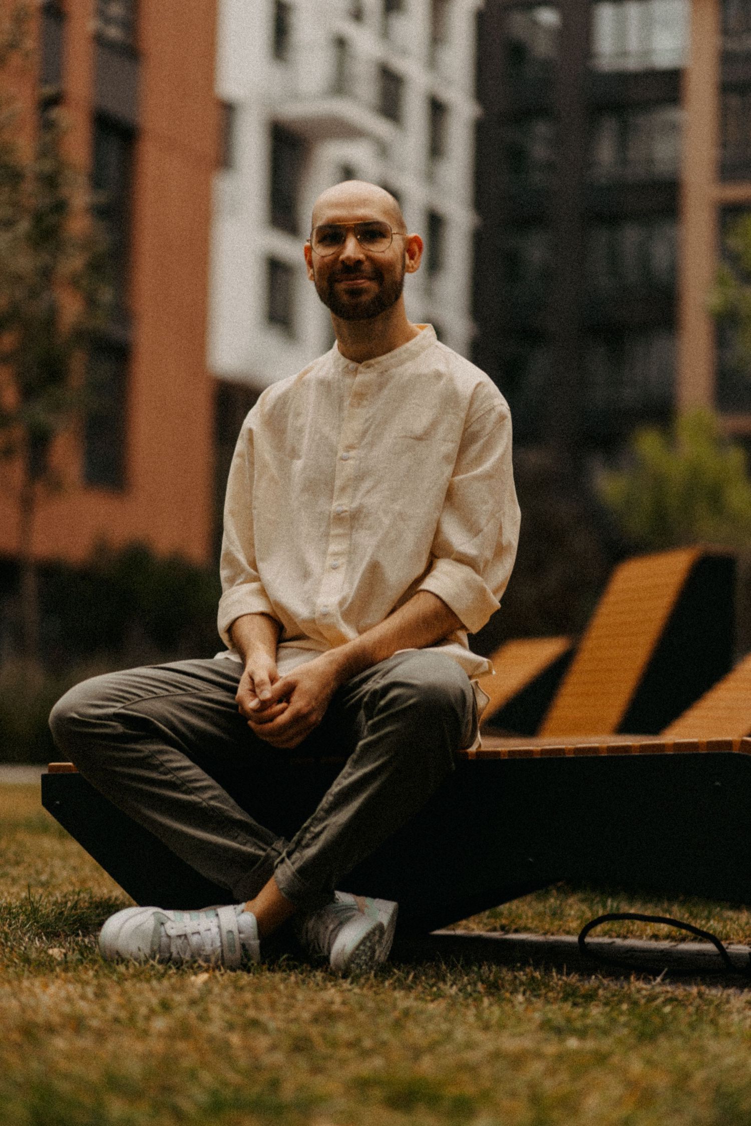 Person in cream colored shirt sits cross-legged on bench in urban garden setting.