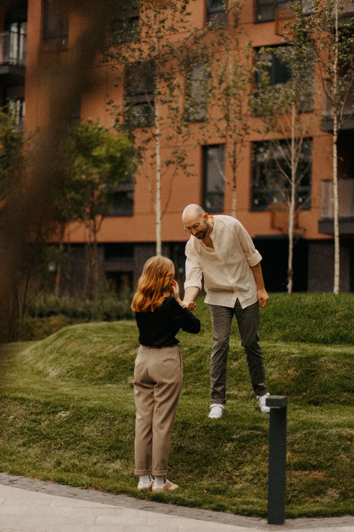 Two people share a playful moment on a grassy hill near an apartment building.
