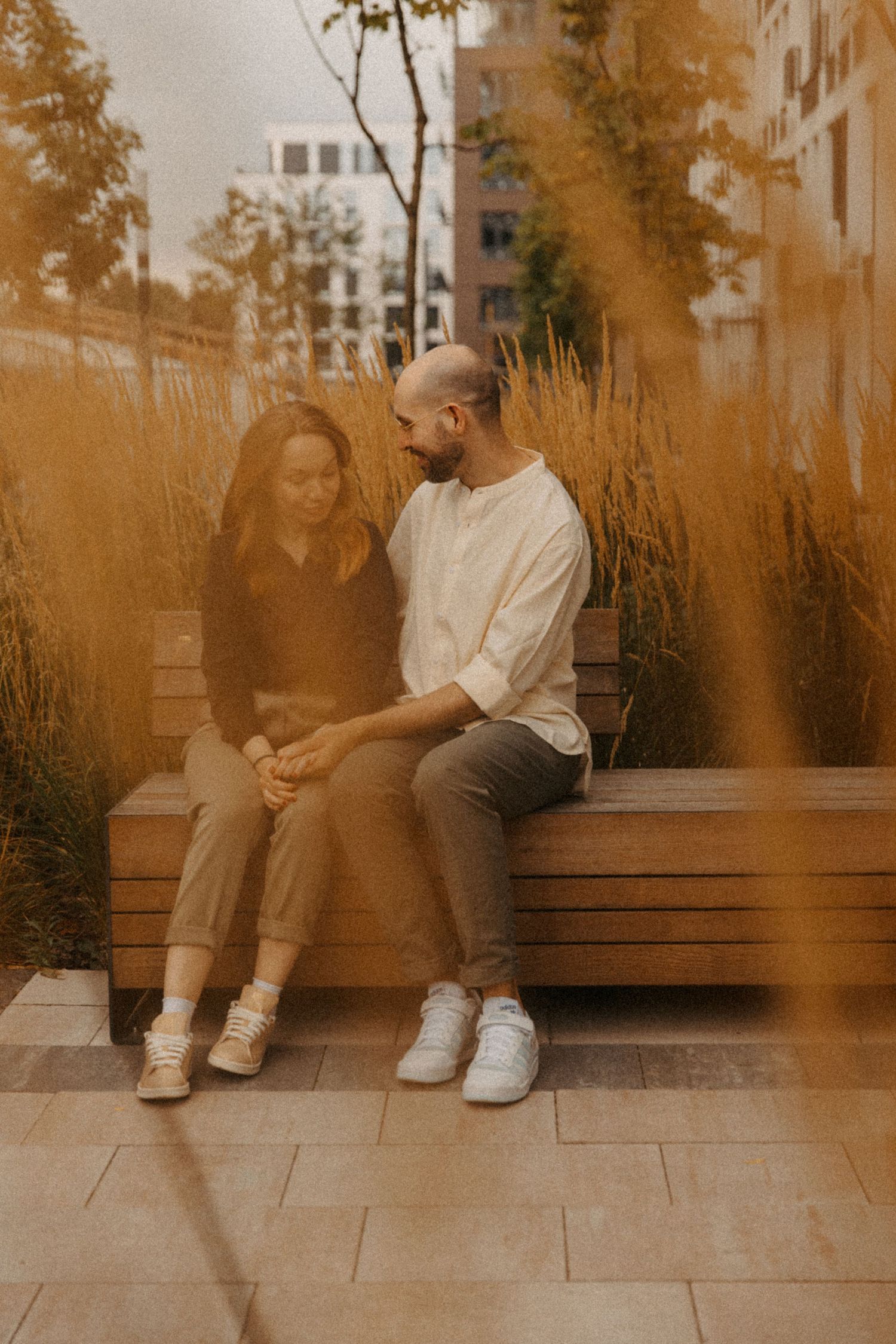 A couple sits together on a wooden bench in an urban setting with autumn colors.