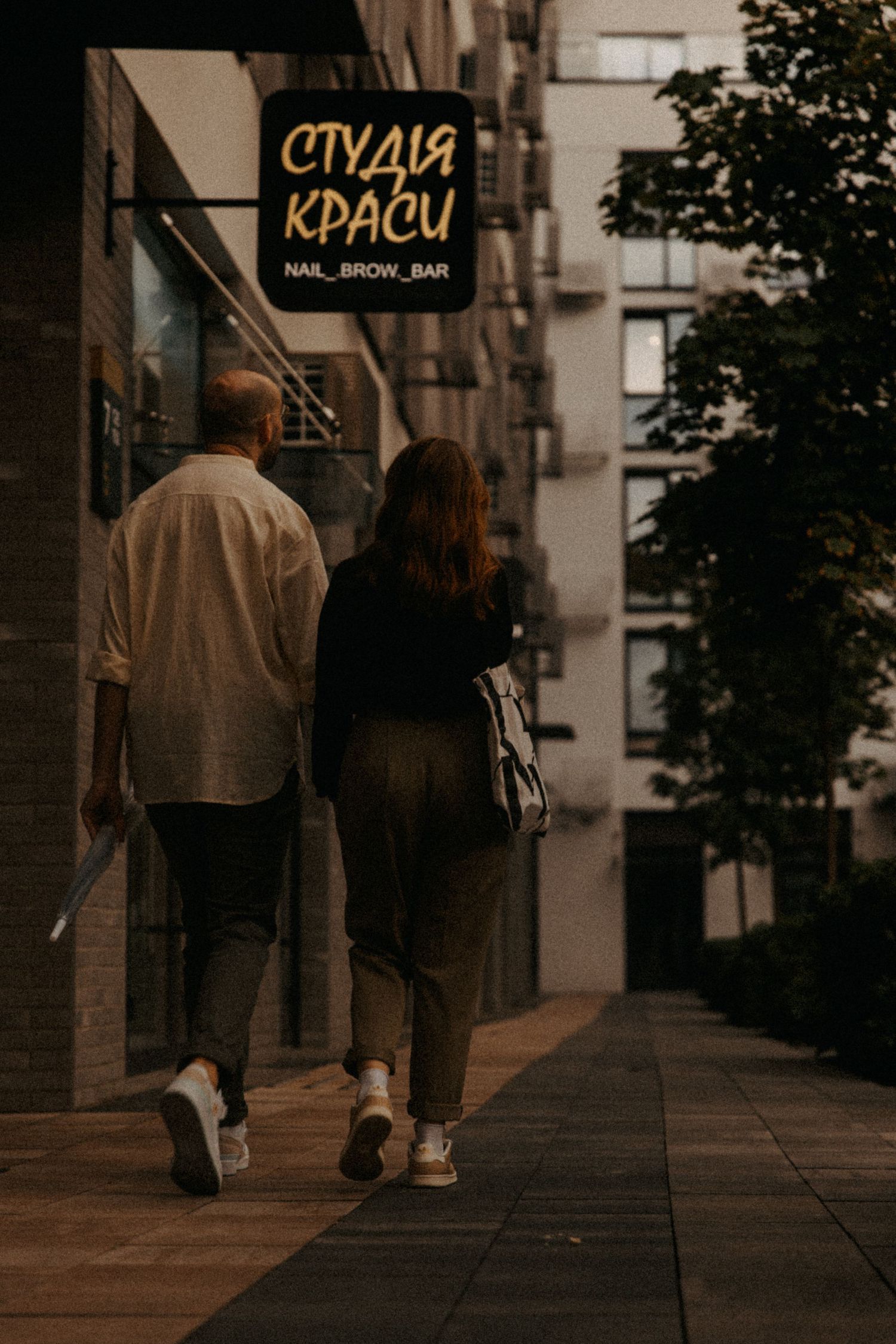 A couple walks together on a city street past a beauty salon sign.