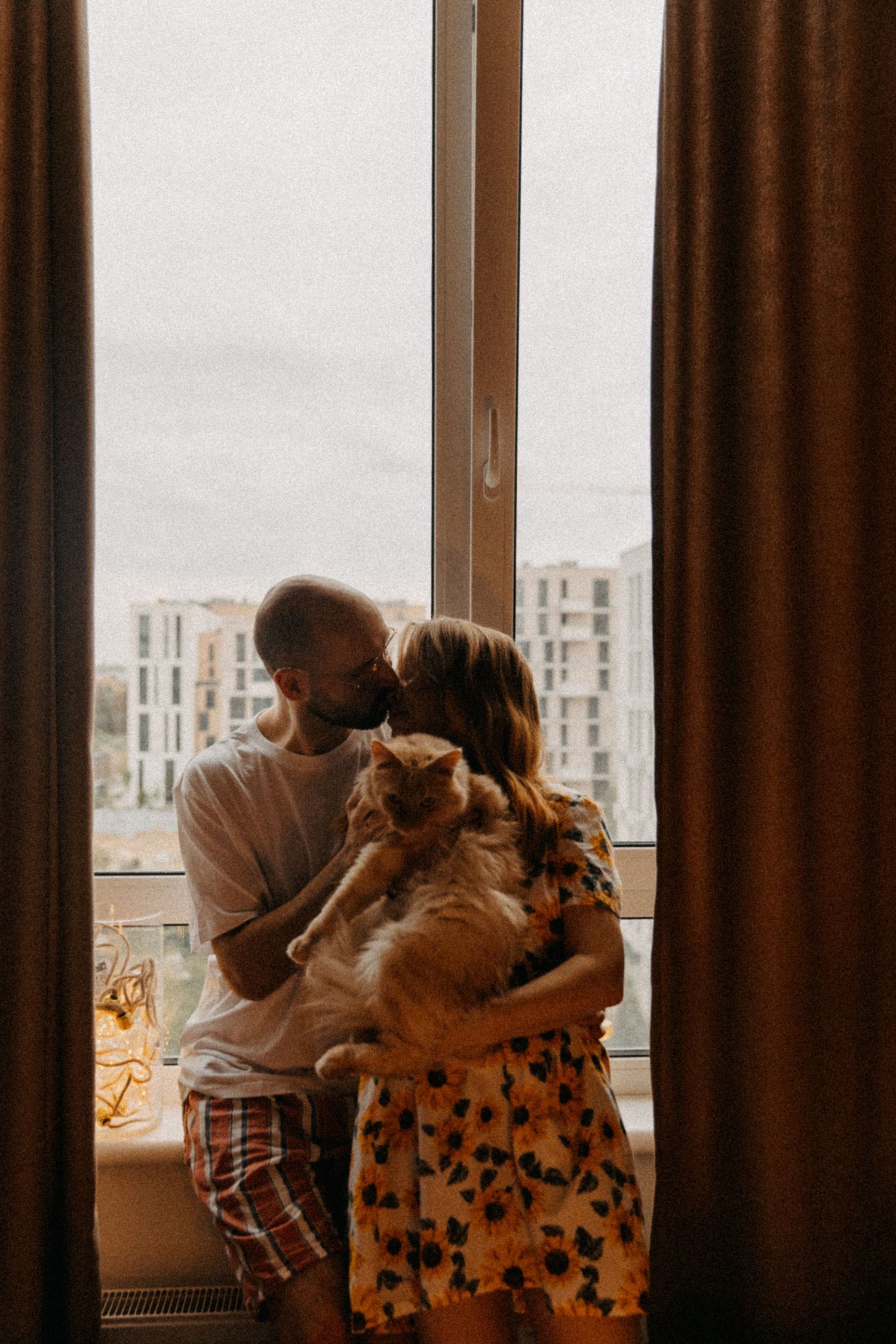 A couple by a window holding their cat with city buildings visible outside.