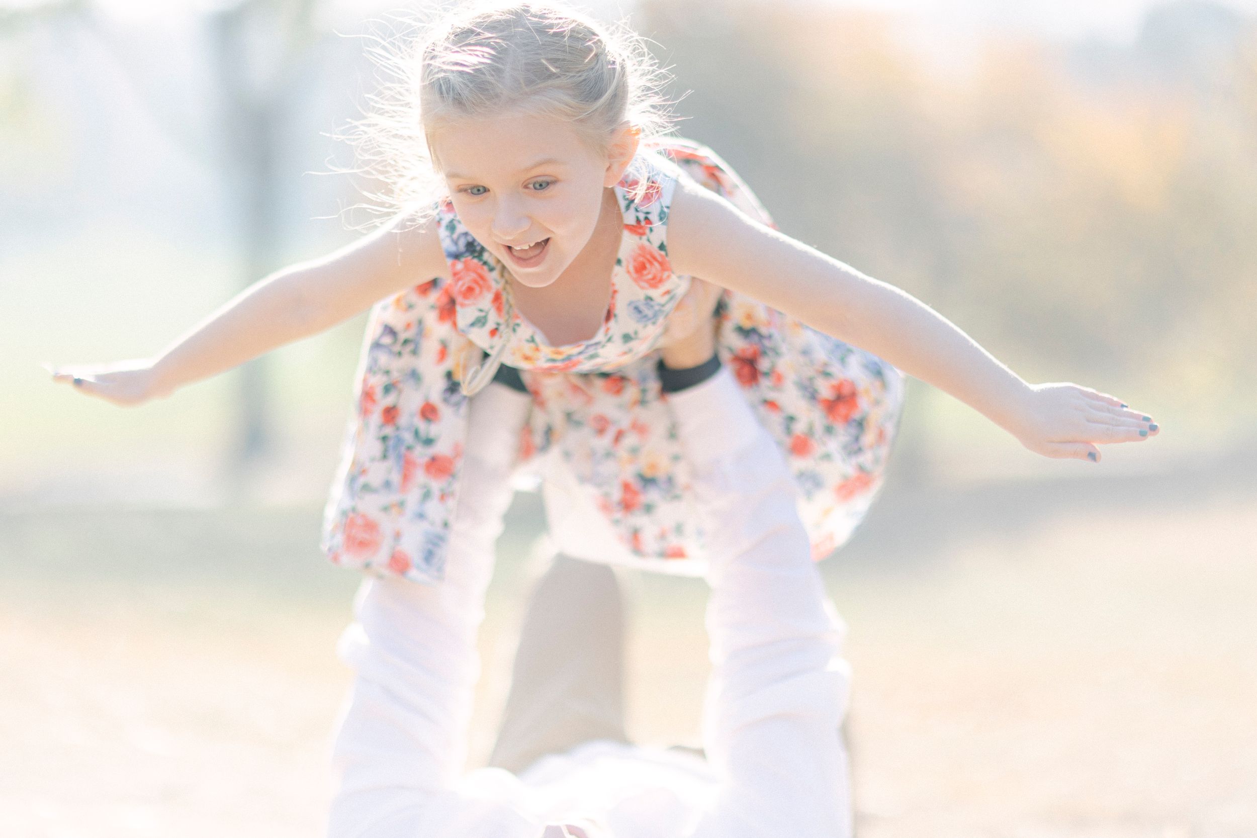 A child in a floral dress spreads their arms like wings while being lifted up outdoors.