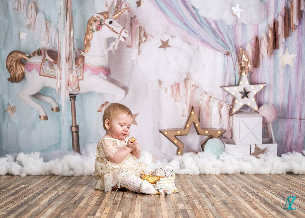 Baby in white dress sits on wooden floor with dreamy carousel and star decorations in soft pastel backdrop.