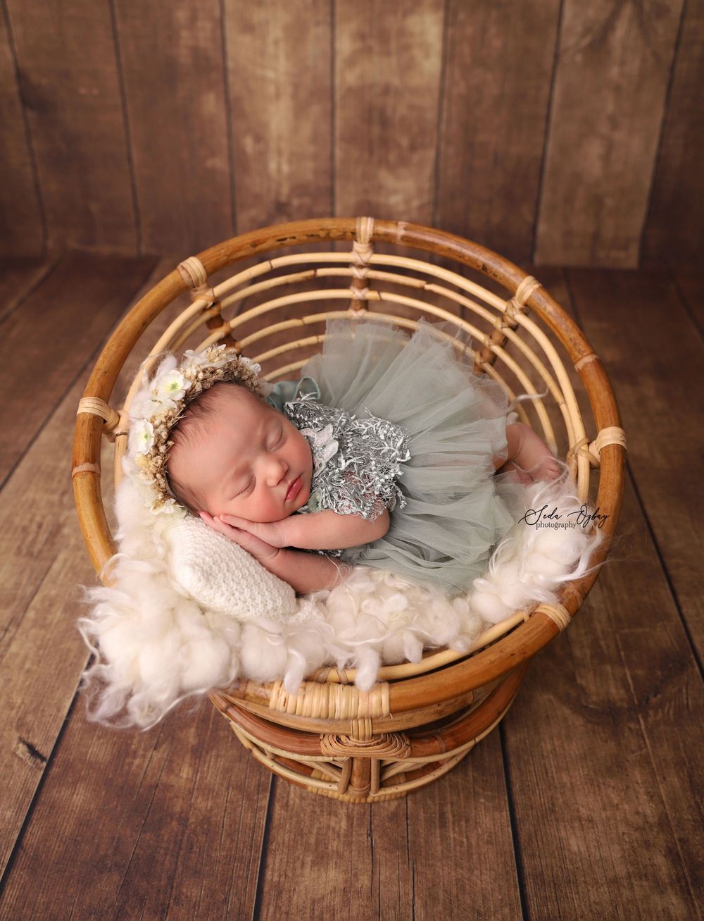 A sleeping newborn in a silver dress nestled in a bamboo basket on wooden floor.