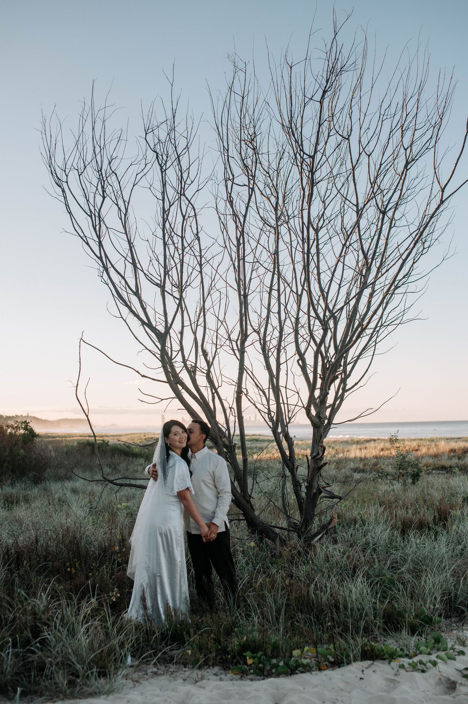 A couple in romantic embrace stands beside a bare tree at dusk with beach and ocean in the background.