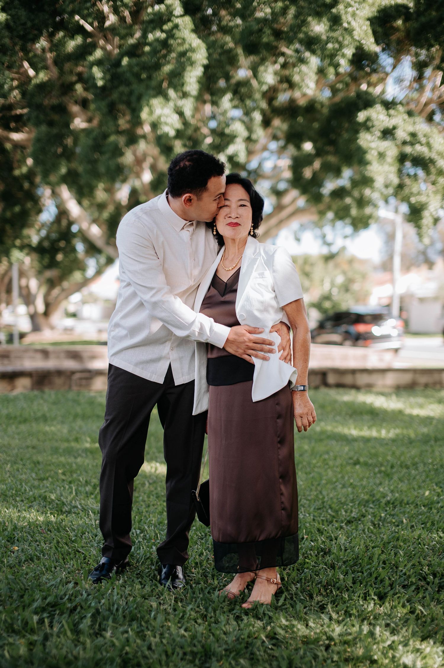 A couple in white and brown outfits share an intimate moment together on a lush green lawn under leafy trees.