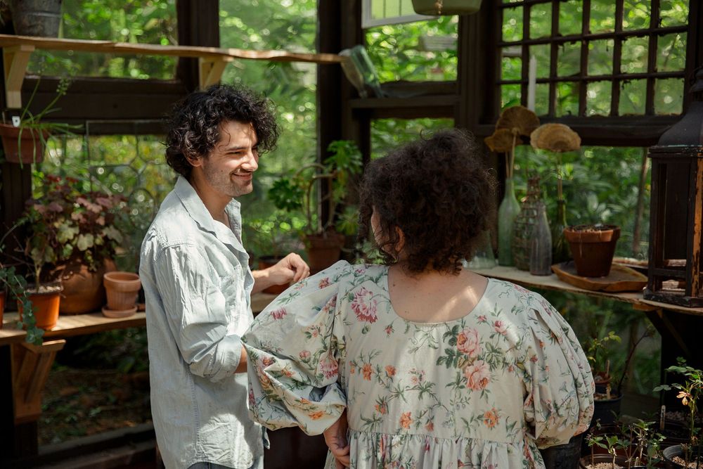 Two people engaged in conversation inside a greenhouse filled with plants and natural light.