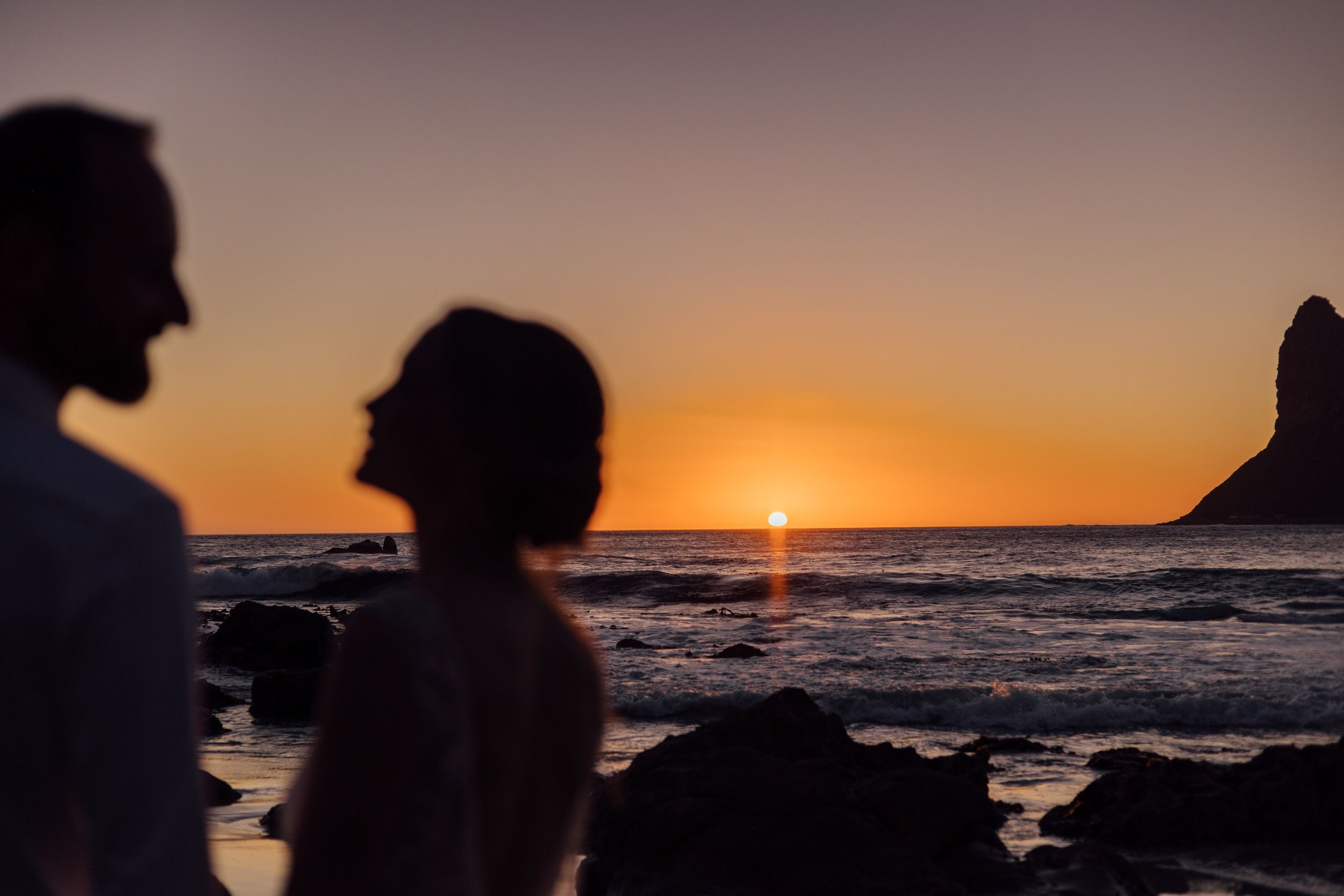 Silhouettes watch a vibrant orange sunset over the ocean with rocks and a sea stack in the distance.