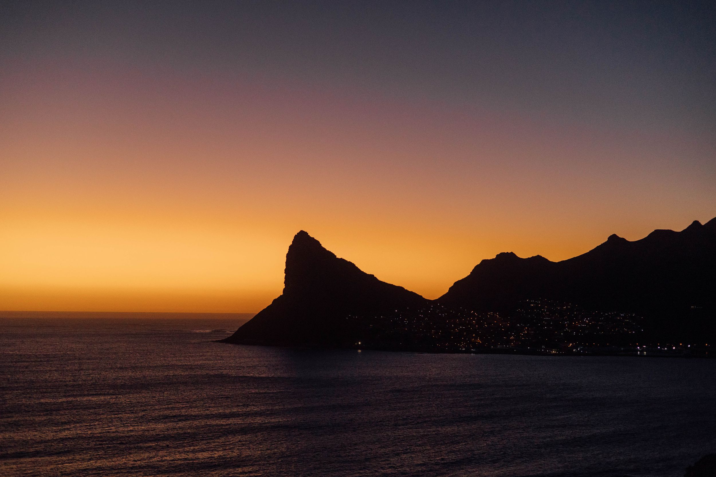 Dramatic silhouette of mountain peaks against an orange and purple sunset sky over calm ocean waters.