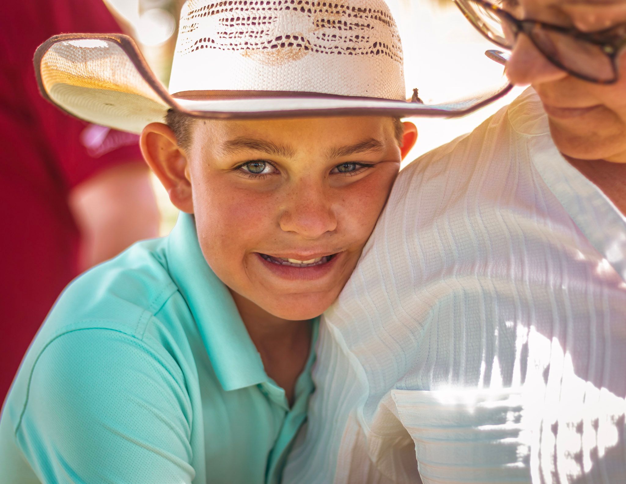 A smiling youth wearing a light cowboy hat and turquoise polo shirt in bright natural lighting.
