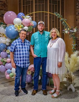 Three people stand together in front of a decorative balloon arch with pastel colors and floral accents.