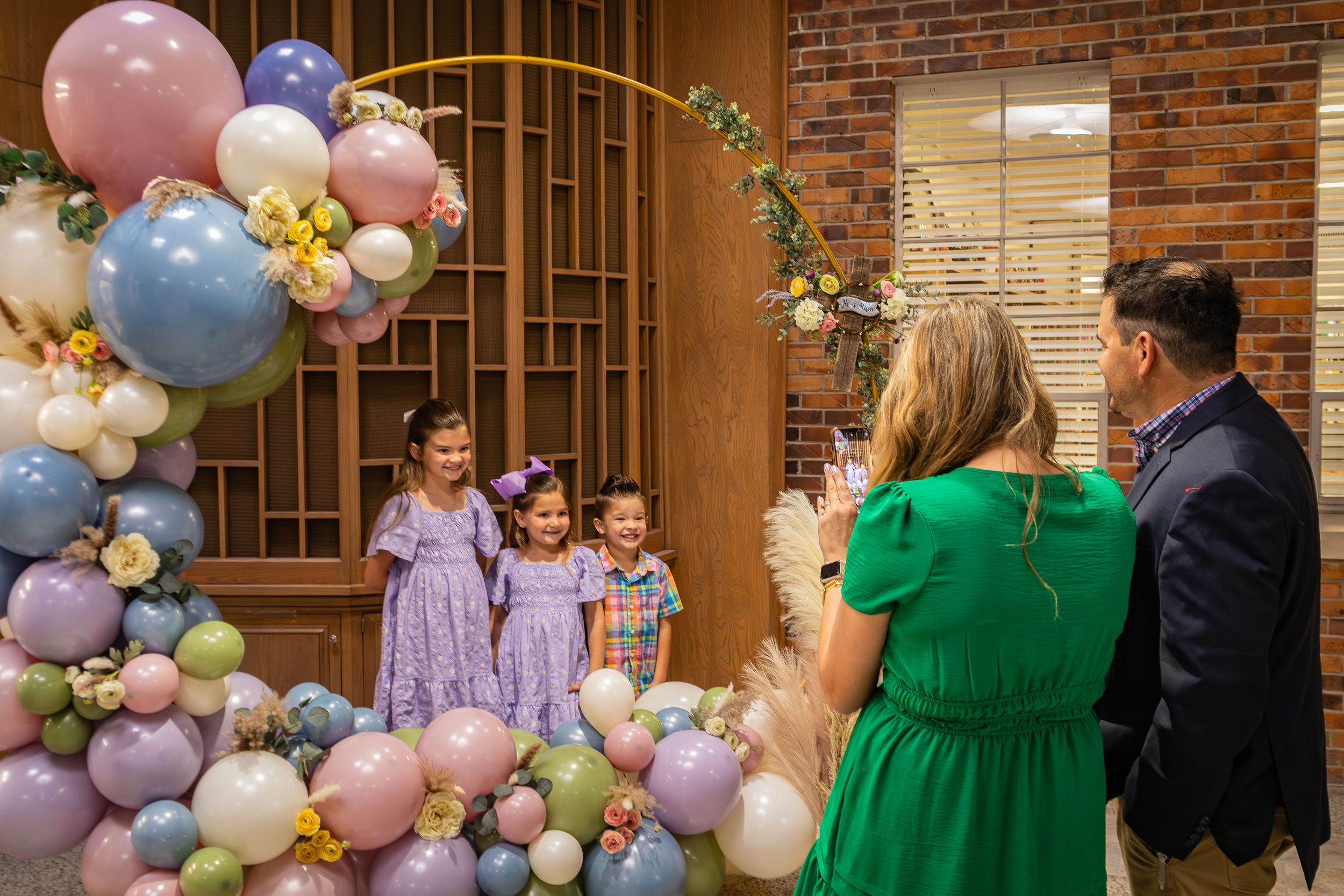 Pastel balloon arch decorates a brick wall entrance with people gathered at a special event in formal attire.