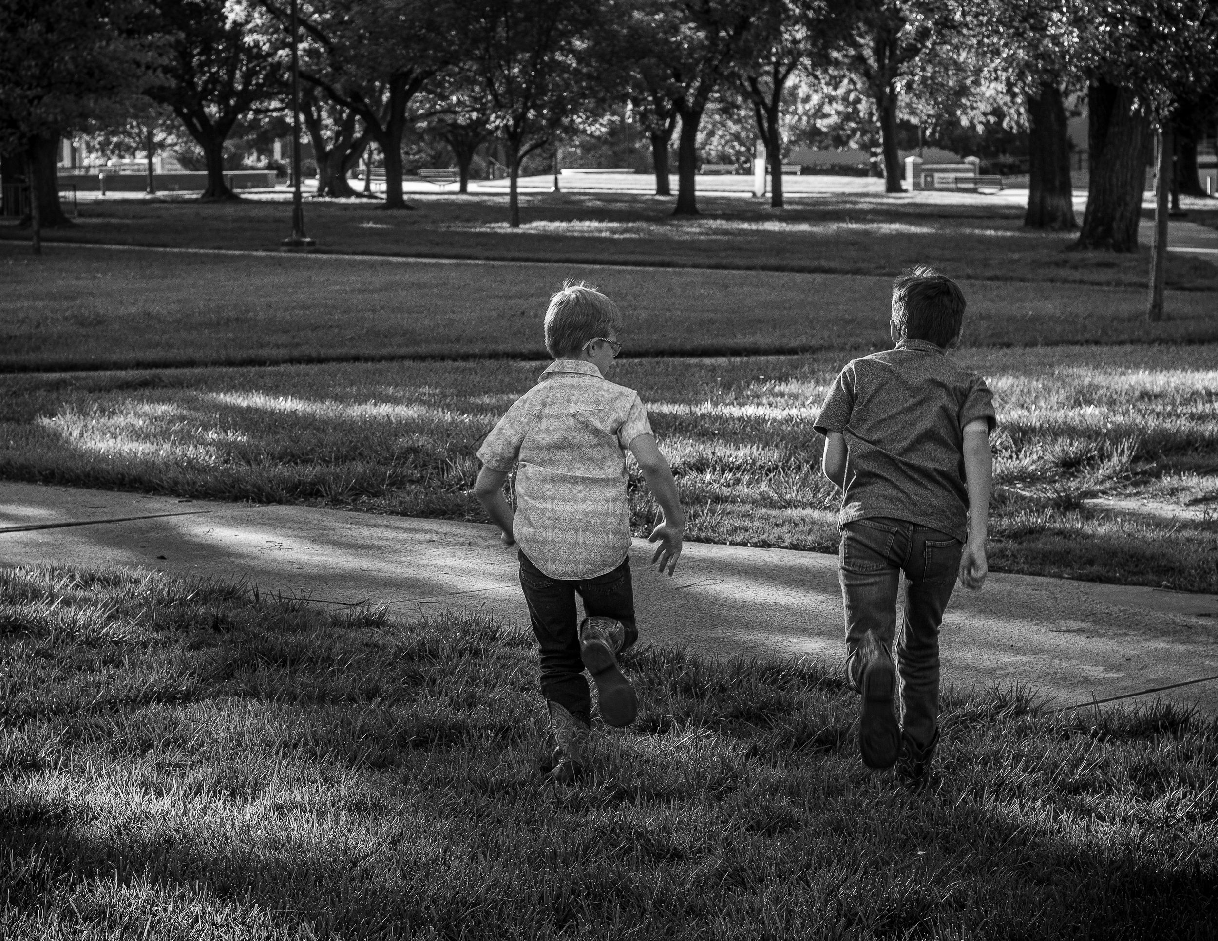 Black and white photo of two children running on a path through a park on a sunny day.