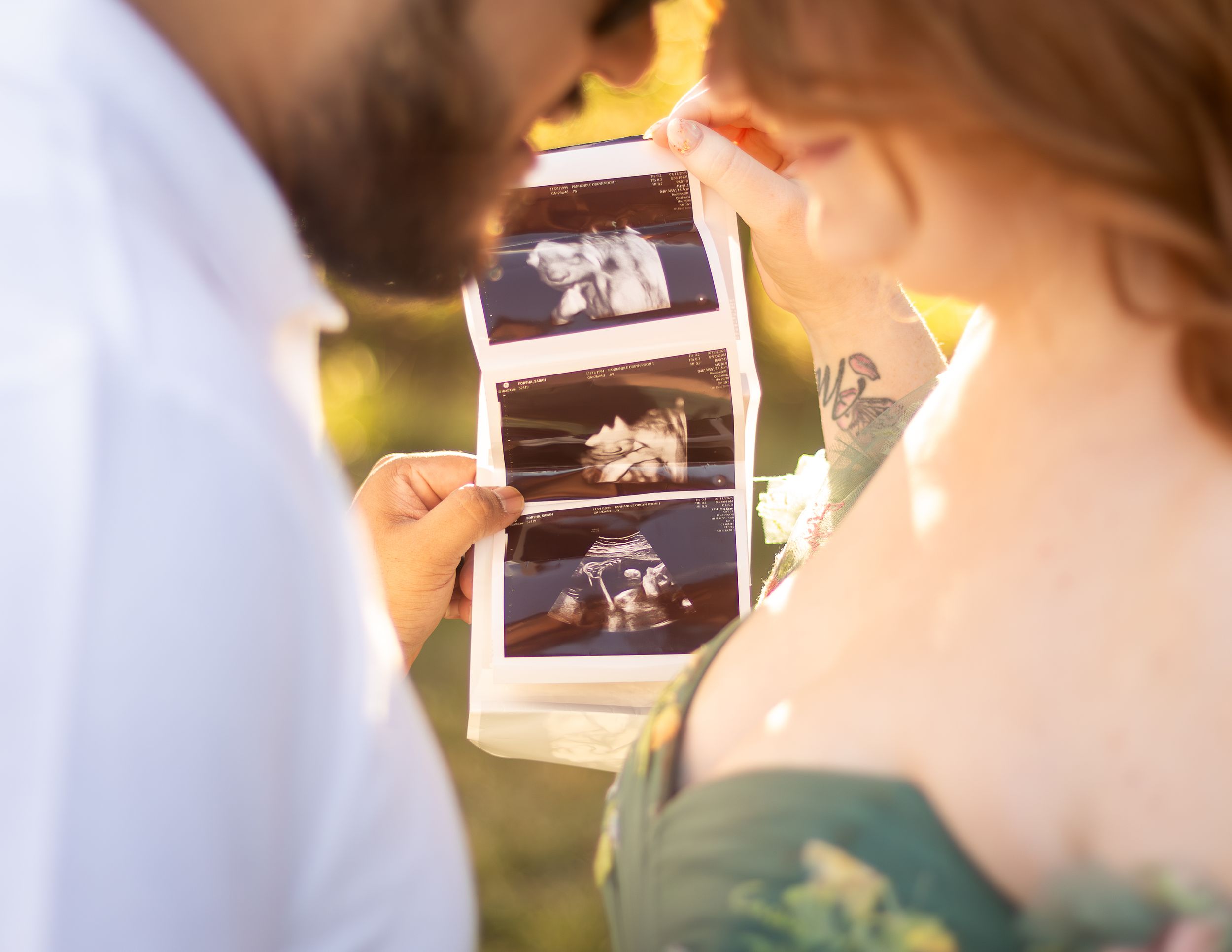 A couple holds ultrasound photos together in soft sunlight while sharing an intimate moment.