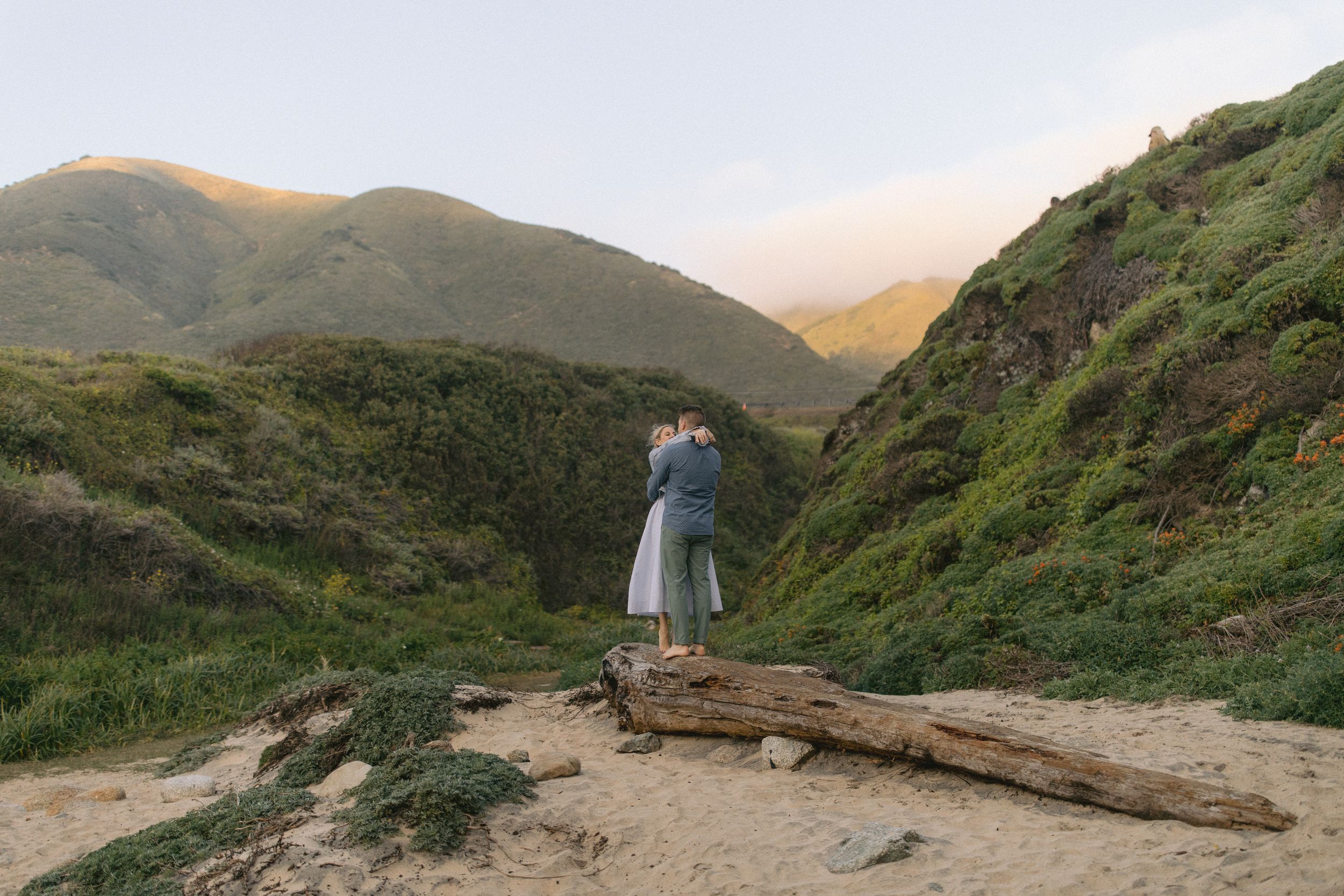 A couple stands on a rugged mountain path at sunset, surrounded by green hills and scenic natural landscape.