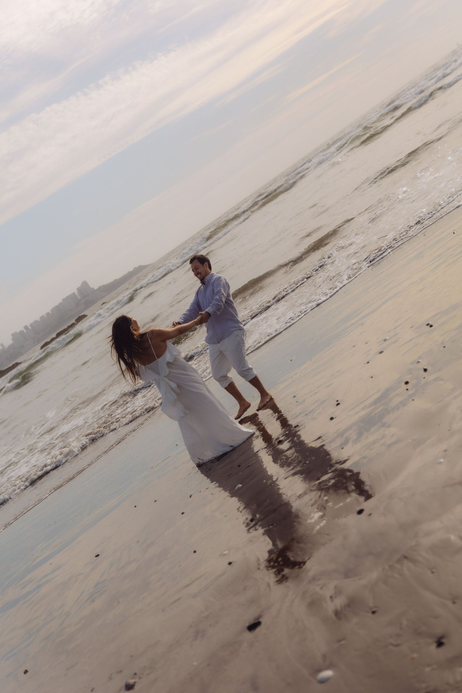 A joyful couple dances and spins together on a sandy beach with gentle waves at sunset.