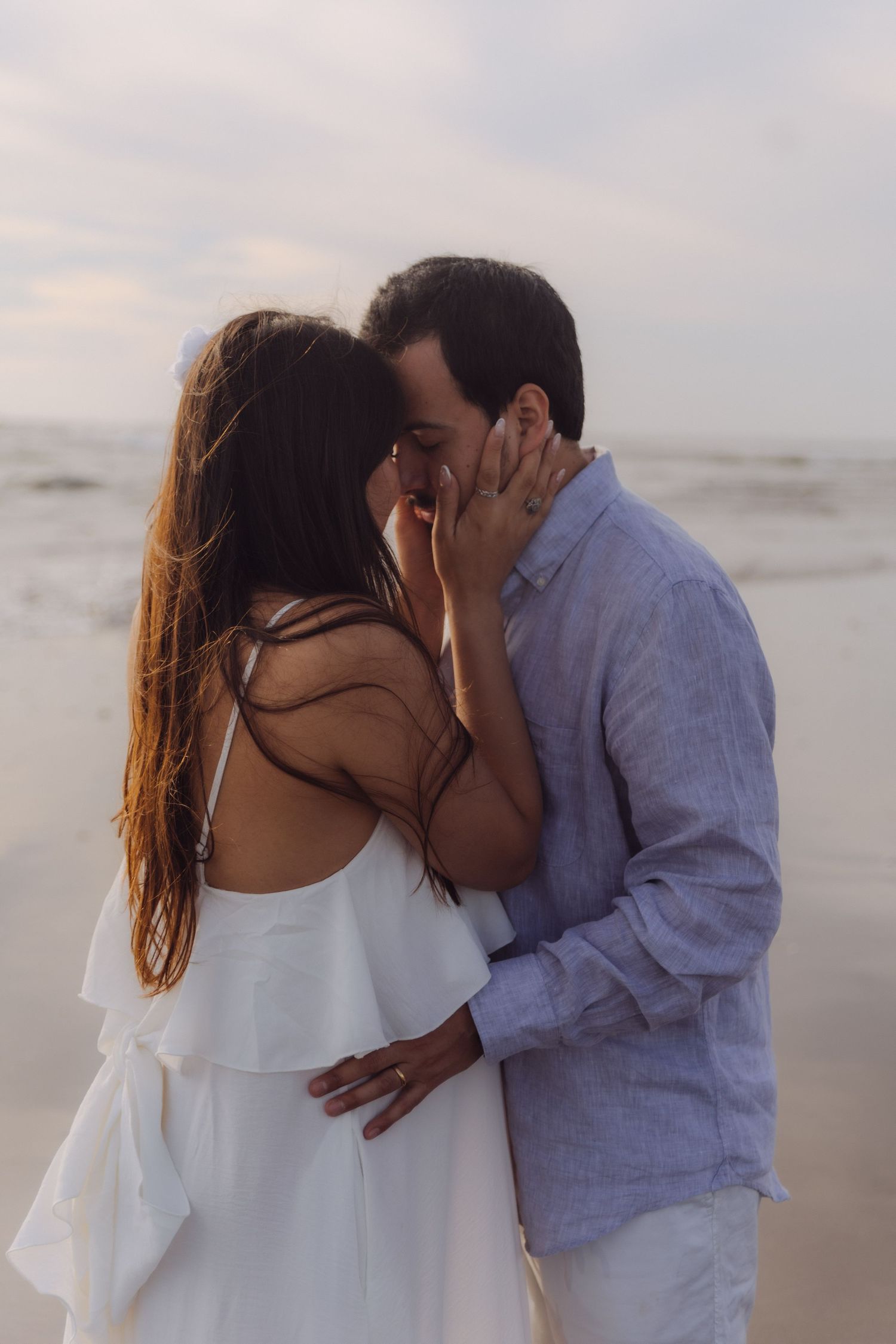 A couple shares a romantic kiss on the beach at dusk, with ocean waves in the background.