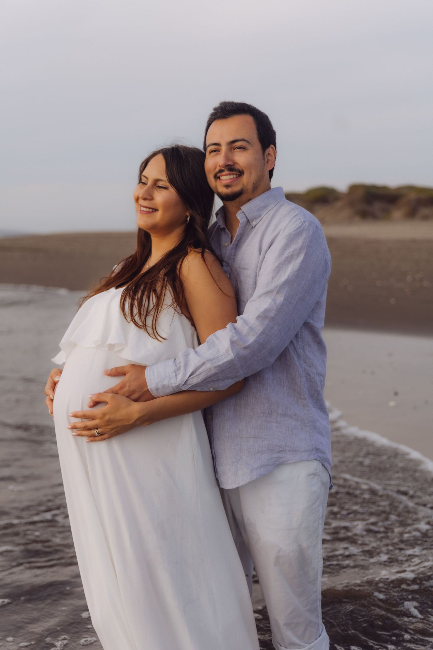 A couple embraces on the beach at sunset, with the man lovingly cradling the womans pregnant belly.