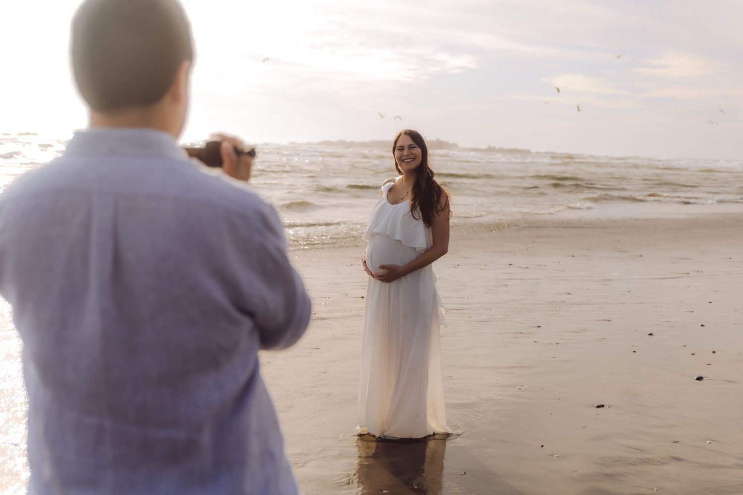 A pregnant woman in a white dress smiles as her partner photographs her on a scenic beach at sunset.