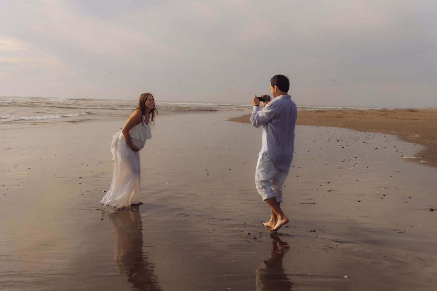 Man photographing pregnant woman in white dress standing in ocean waves on a beach at sunset.