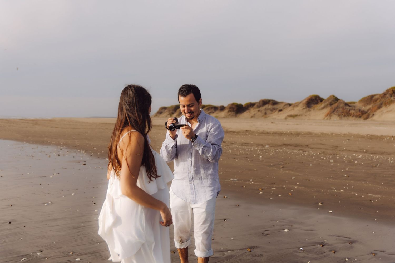 A couple enjoys a playful moment on a sandy beach, with the man photographing his partner at golden hour.