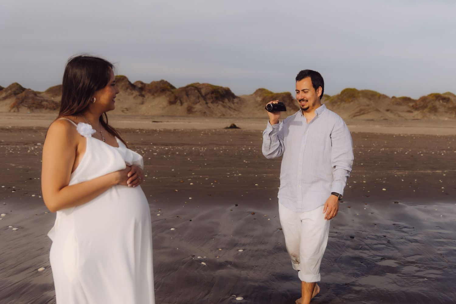 Happy couple on beach, man smiling at pregnant woman in white dress near sandy shoreline.