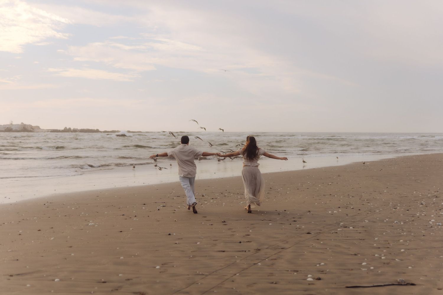 Couple running hand in hand along sandy beach shoreline with seagulls in cloudy sky.
