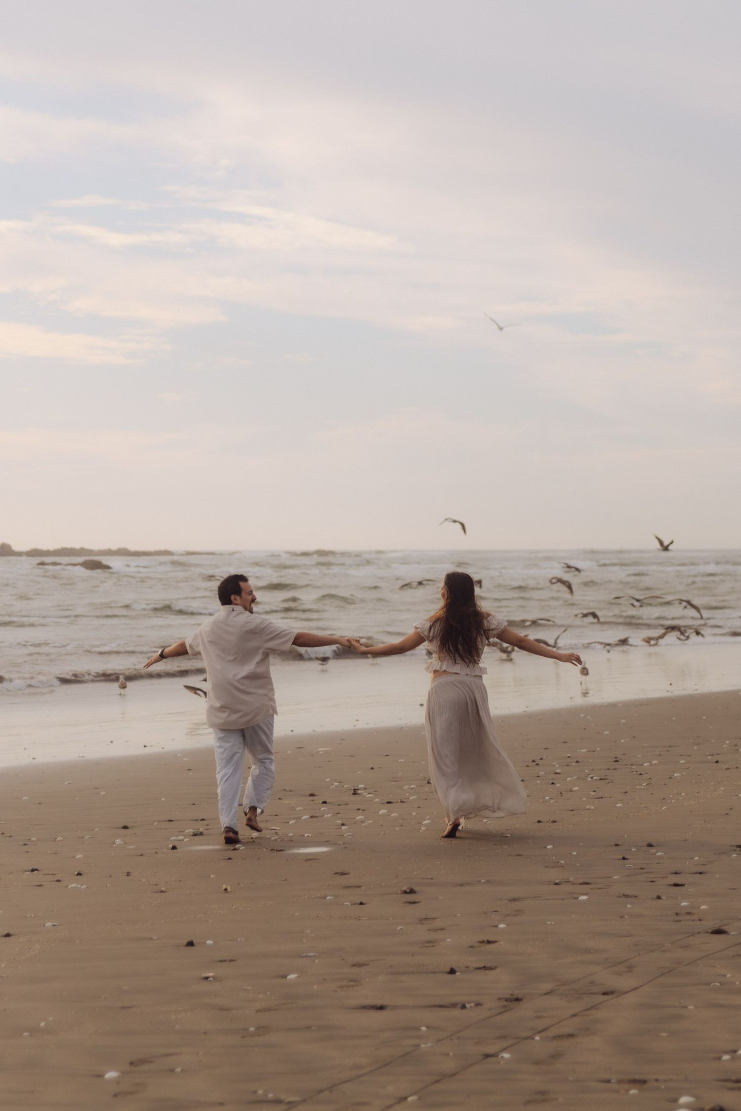 Couple holding hands dancing on beach with seagulls flying overhead near ocean waves.
