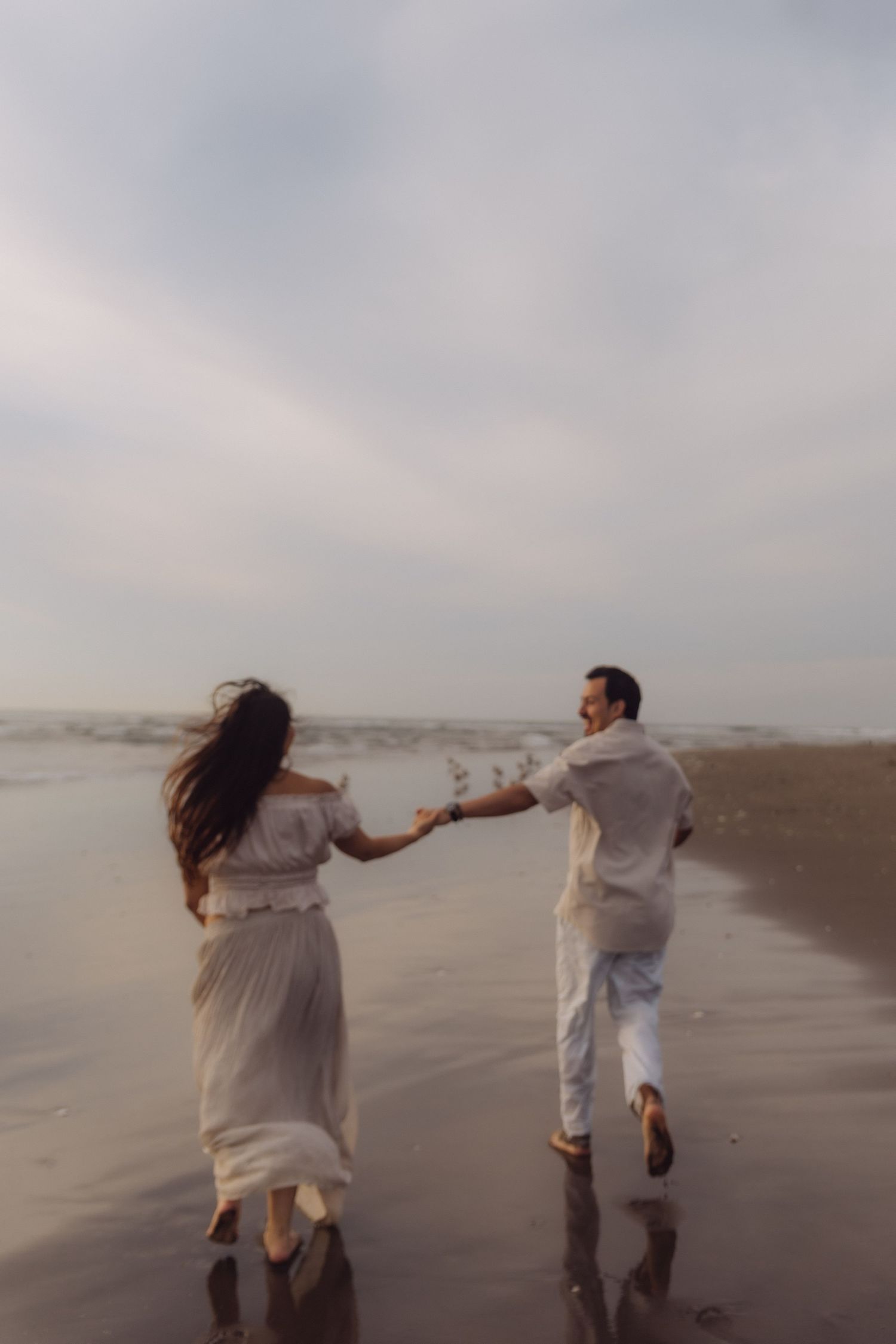 Couple in white outfits holding hands running along wet beach shore with ocean waves.